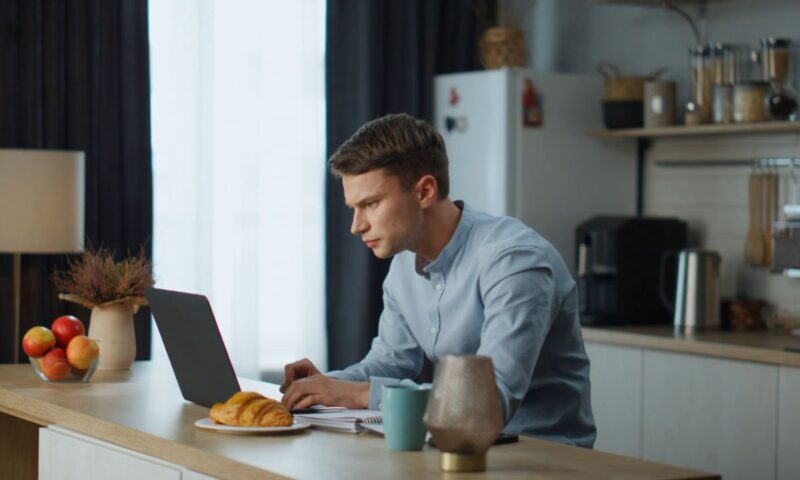momak radi za laptopom od kuće, pored njega kafa, knjiga i kroasan
